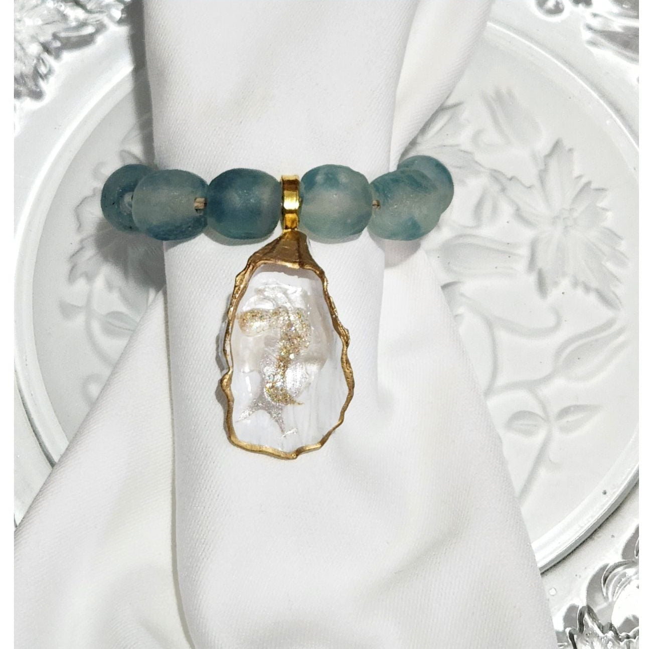 Hand-painted oyster shell napkin ring featuring deep Blue Wave Marine recycled glass beads and a gold wood accent on a while linen napkin
