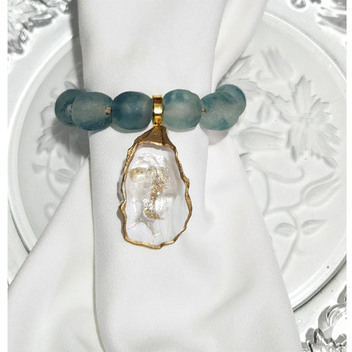 Hand-painted oyster shell napkin ring featuring deep Blue Wave Marine recycled glass beads and a gold wood accent on a while linen napkin
