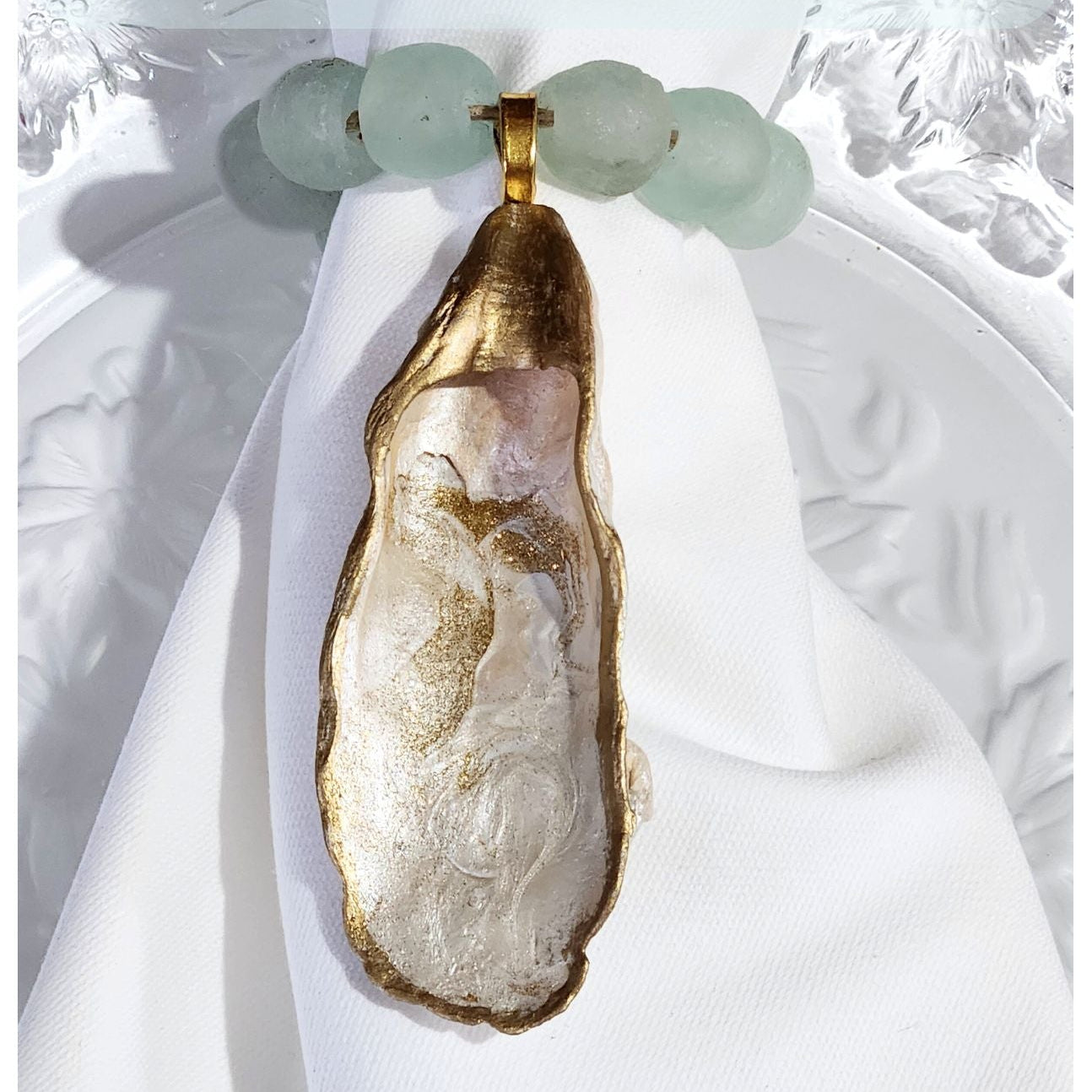 Hand-painted, gilded oyster shell napkin ring featuring translucent aqua recycled glass beads on a white linen napkin