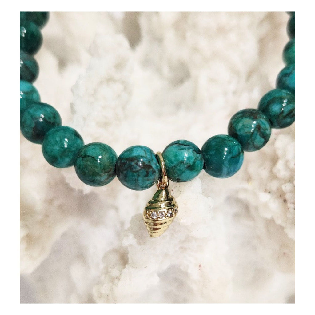 Close up shot of the Deep Sea African Turquoise stretch bracelet featuring 6mm green gemstone beads and a gilded conch shell charm with micro CZ accents displayed on a white coral reef decor piece by Coastline Boutique