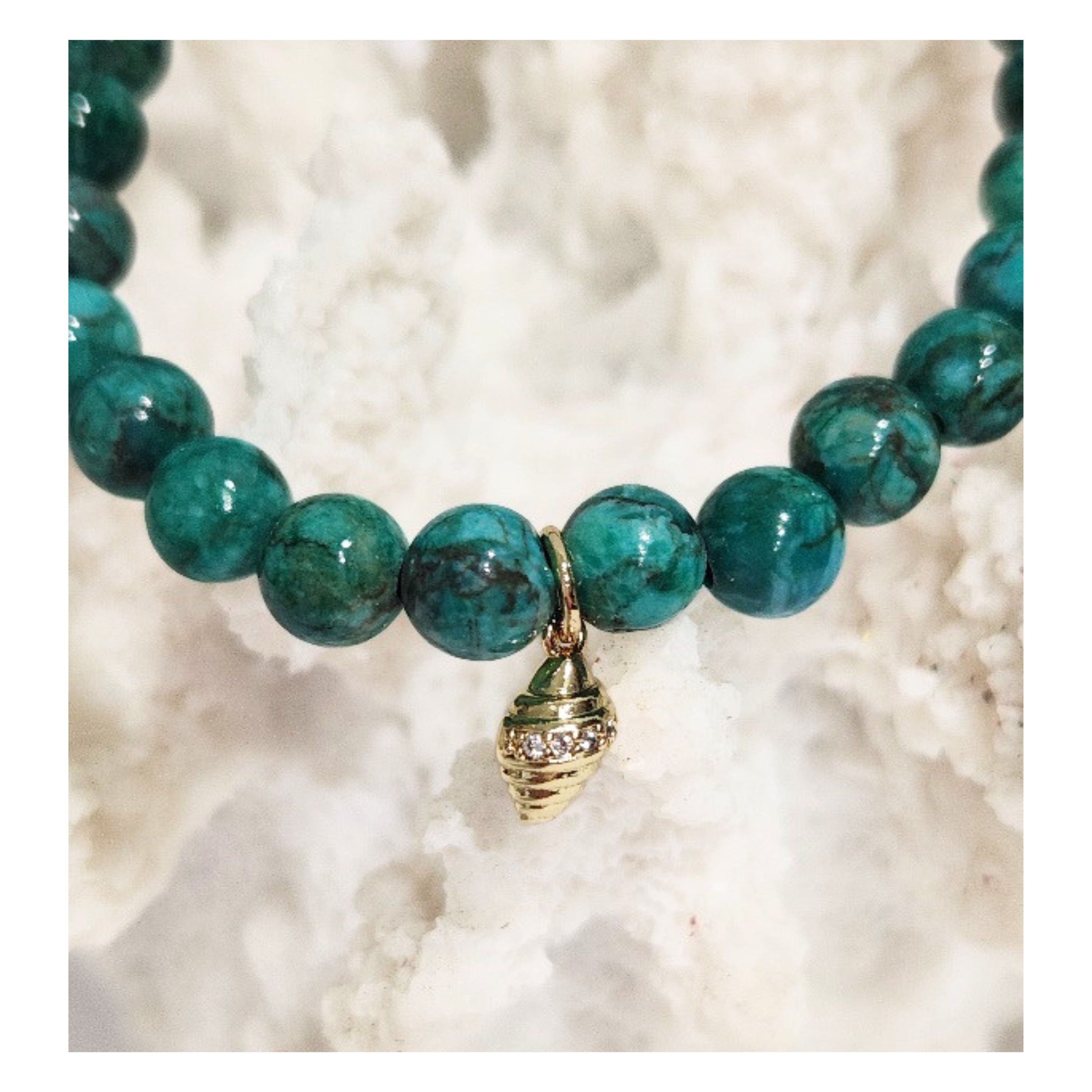Close up shot of the Deep Sea African Turquoise stretch bracelet featuring 6mm green gemstone beads and a gilded conch shell charm with micro CZ accents displayed on a white coral reef decor piece by Coastline Boutique