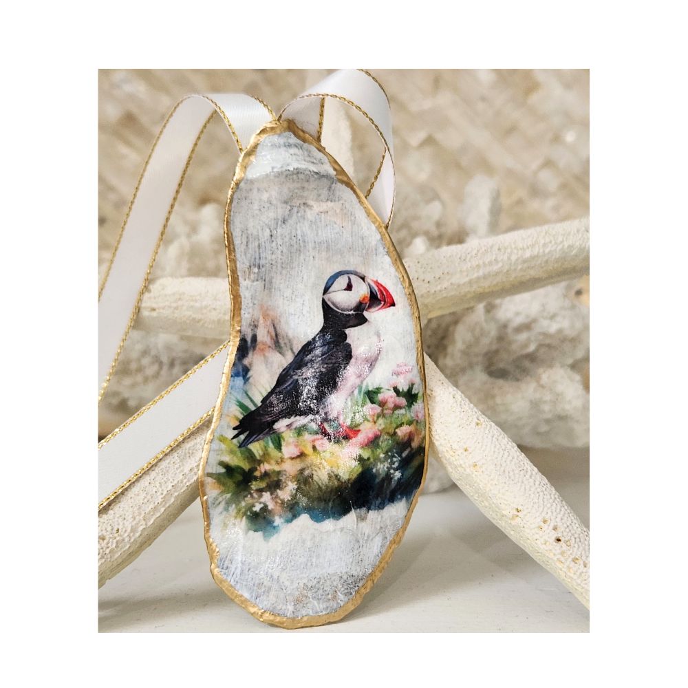 A handcrafted gilded oyster shell ornament featuring a detailed decoupaged Atlantic Puffin design. The puffin is depicted sitting amidst wild grass and tiny flowers. The ornament includes a white and gold satin ribbon hanger and is displayed against a natural starfish and white coral