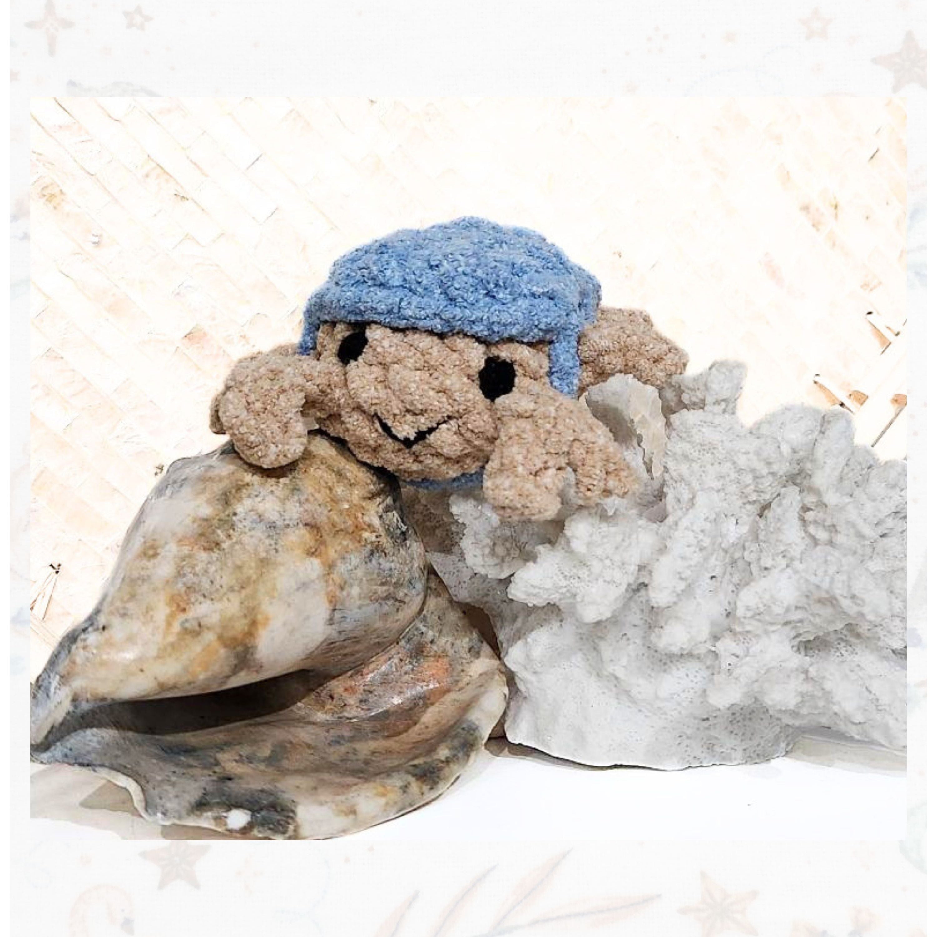 Hand-crocheted Brejoy Crabulous pocket companion in a neutral sand color with a bright blue artisan sun hat. The minature crab is perched atop a large, textured shell and a piece of white reef, showcasing its palm-sized scale and soulful hand-embroidered eyes in a coastal setting.