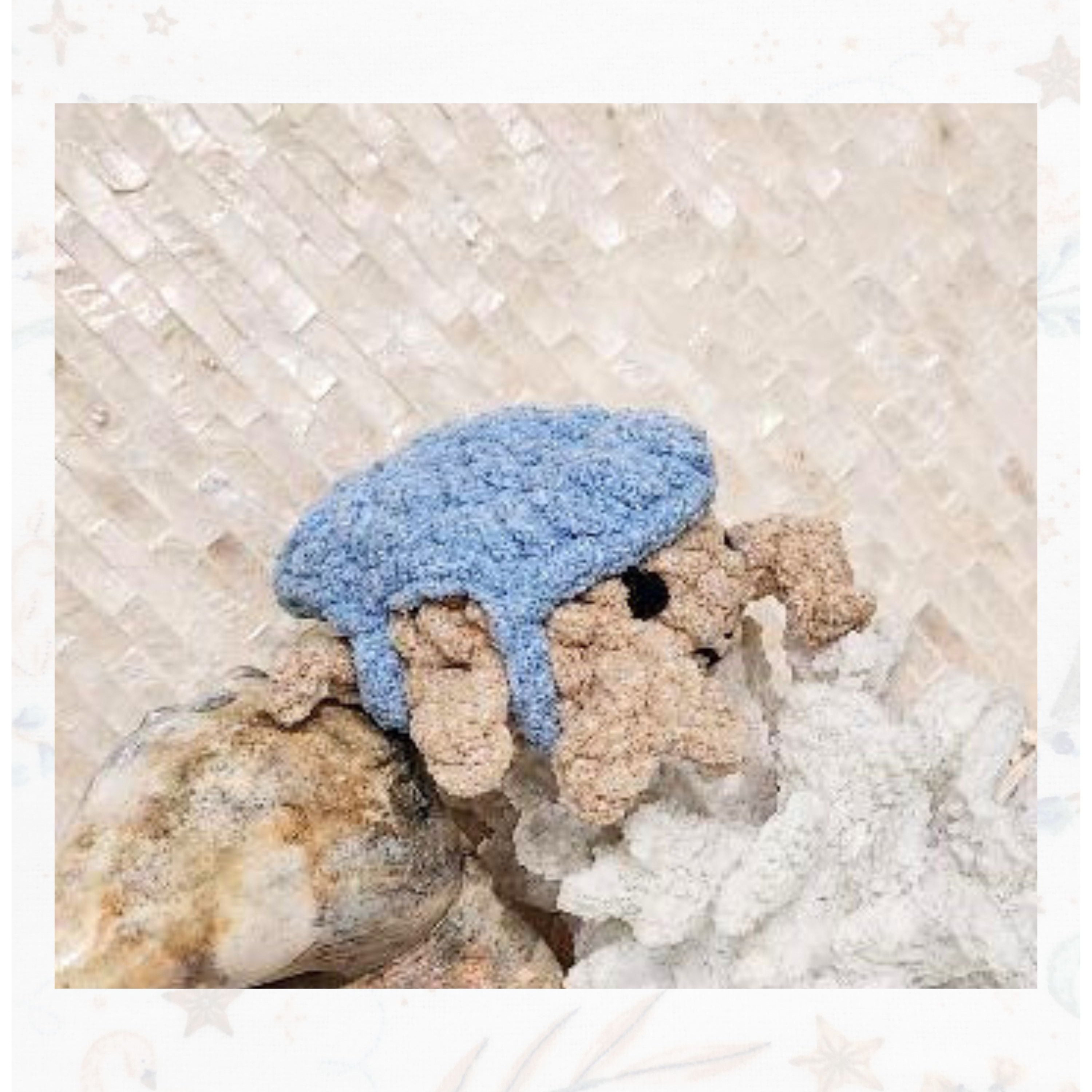 Side view of Crabulous the sand-toned crochet crab scuttling across a large shell toward a white coral reef decor piece by Coastline Boutique