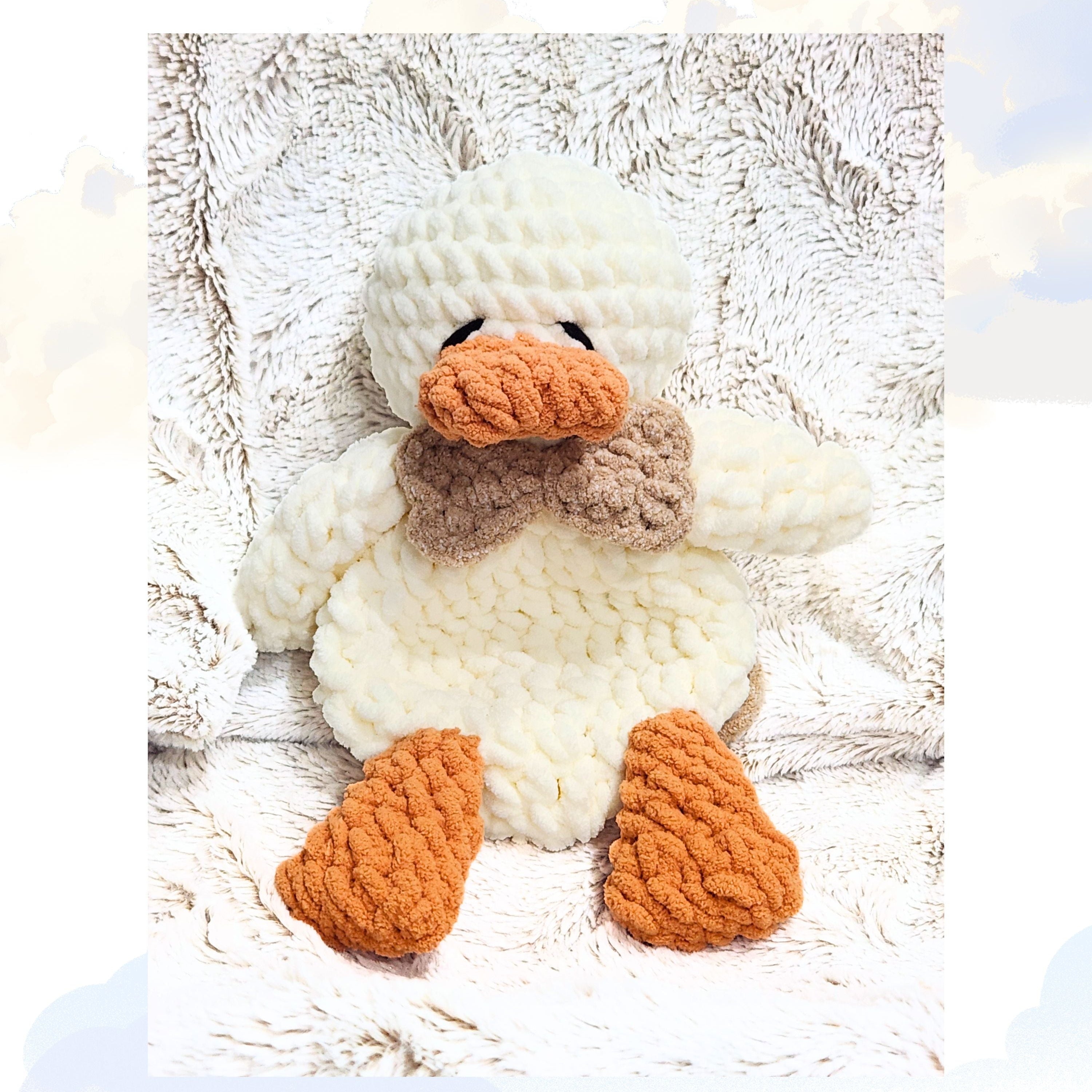 A Ducky Dreams vanilla cream crochet duck snuggler wearing a dapper driftwood brown bow tie.  The duck is shown in a full body view, laying peacefully on a plush blanket with a whimsical, soft focus background of clouds