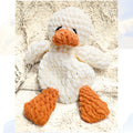 The classic Ducky Dreams vanilla cream crochet duck snuggler in a full body view. The plush duck is nestled on a soft blanket against a whimsical background of distant clouds, showcasing its simple, elegant design and safety first embroidered eyes