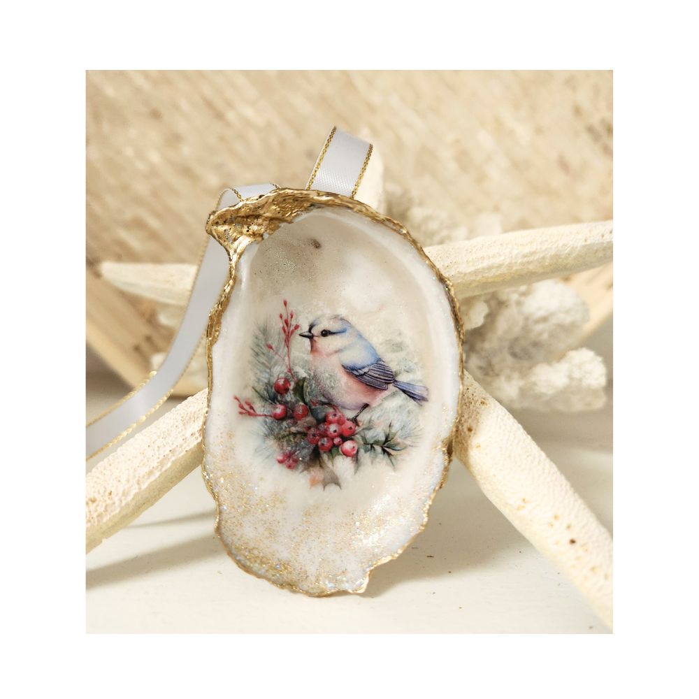 A handcrafted gilded oyster shell ornament featuring a decoupaged Eastern Bluebird perched on a twig with a small fern and red berries. The inside of the shell features a delicate touch of sparkle, finished with a white and gold satin ribbon hanger against a starfish and white coral,