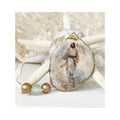 A handcrafted gilded oyster shell ornament featuring a serene design of Jesus walking along the sand, decoupaged with fine rice paper. The hanger is adorned with a clear aqua recycled glass bead flanked by two gold wooden beads, finished with a crisp white and gold trimmed ribbon against a starfish and white coral background