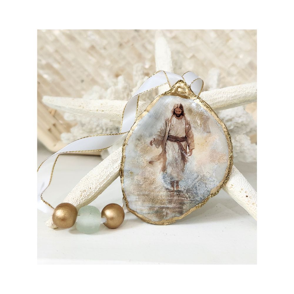 A handcrafted gilded oyster shell ornament featuring a serene design of Jesus walking along the sand, decoupaged with fine rice paper. The hanger is adorned with a clear aqua recycled glass bead flanked by two gold wooden beads, finished with a crisp white and gold trimmed ribbon against a starfish and white coral background