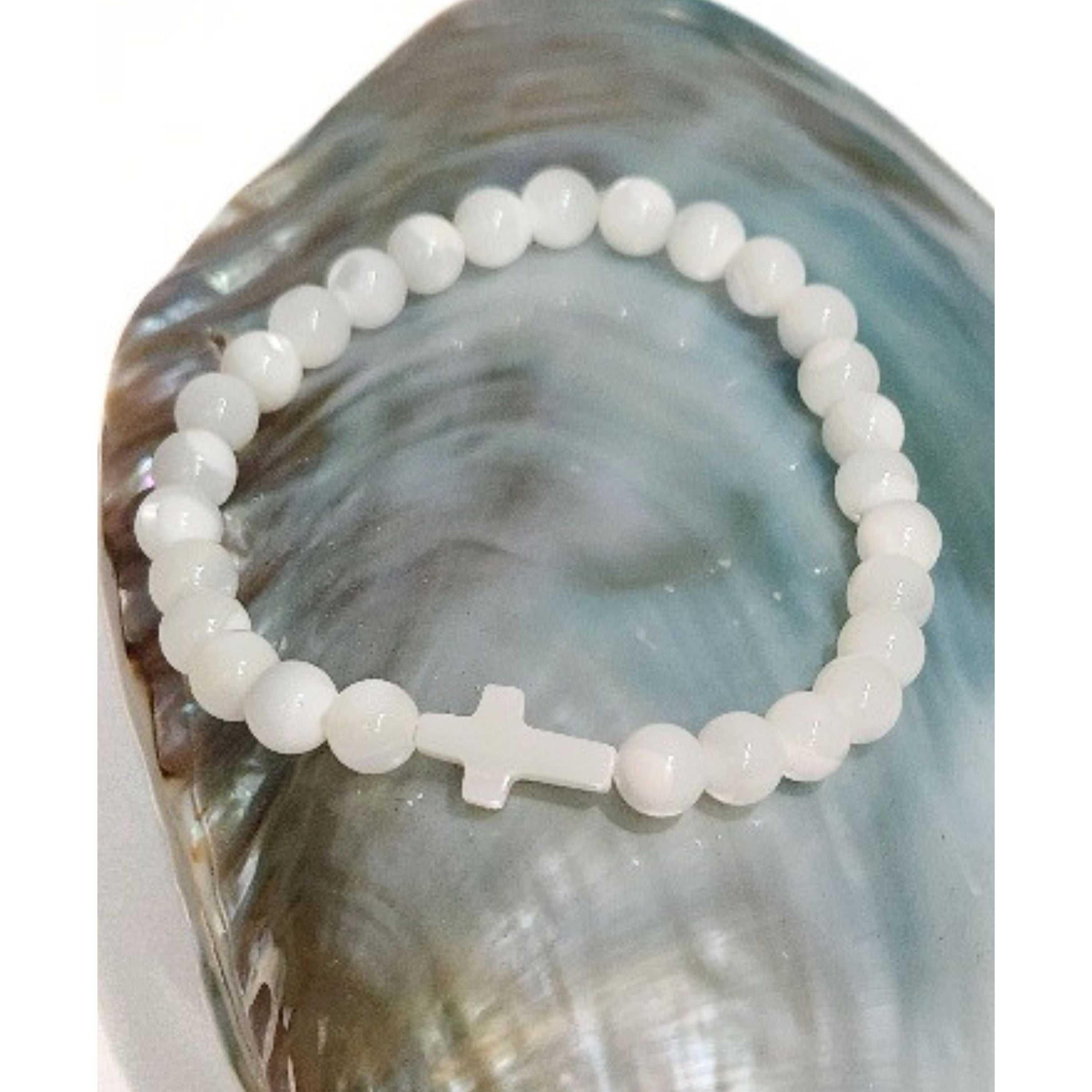 Close up of a Mother of Pearl cross stretch bracelet resting on an iridescent blue shell against a clean white background