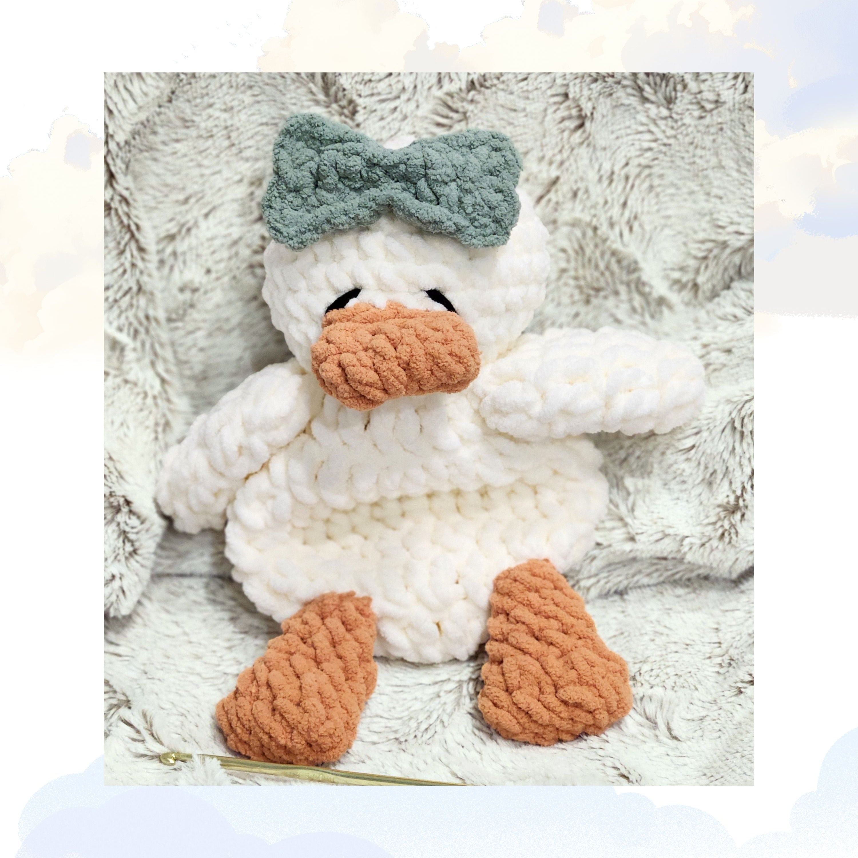 A Ducky Dreams vanilla cream plush duck snuggler featuring a meadow sage green head bow.  The duck is nestled cozily in a plush blanket, set against a whimsical soft background of fluffy clouds to create a dreamy ethereal atmosphere.