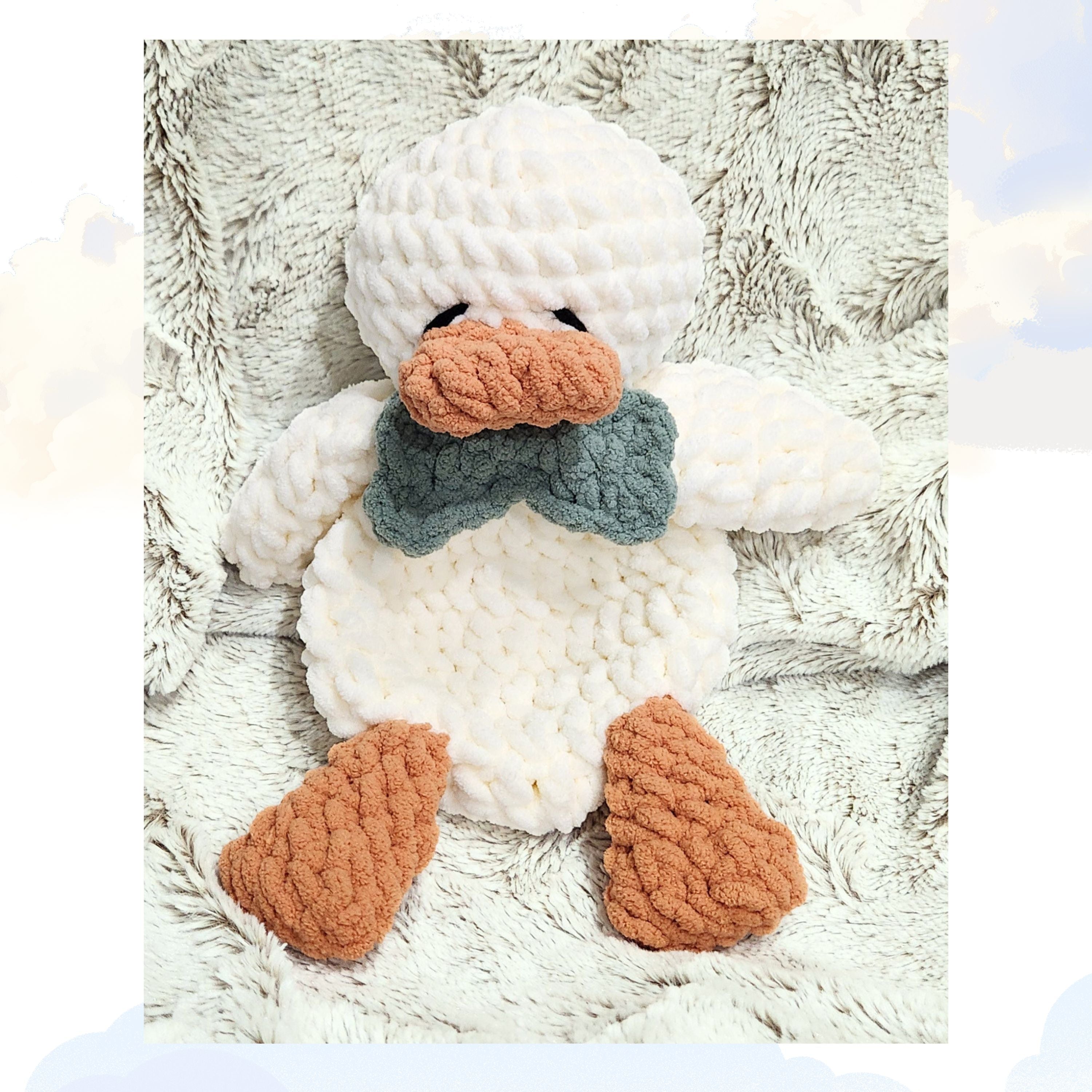 A Ducky Dreams vanilla cream duck snuggler featuring a dapper meadow sage green bow tie. The duck is shown in a full body view, laying cozily on a plush blanket with a dreamy background of whimsical clouds