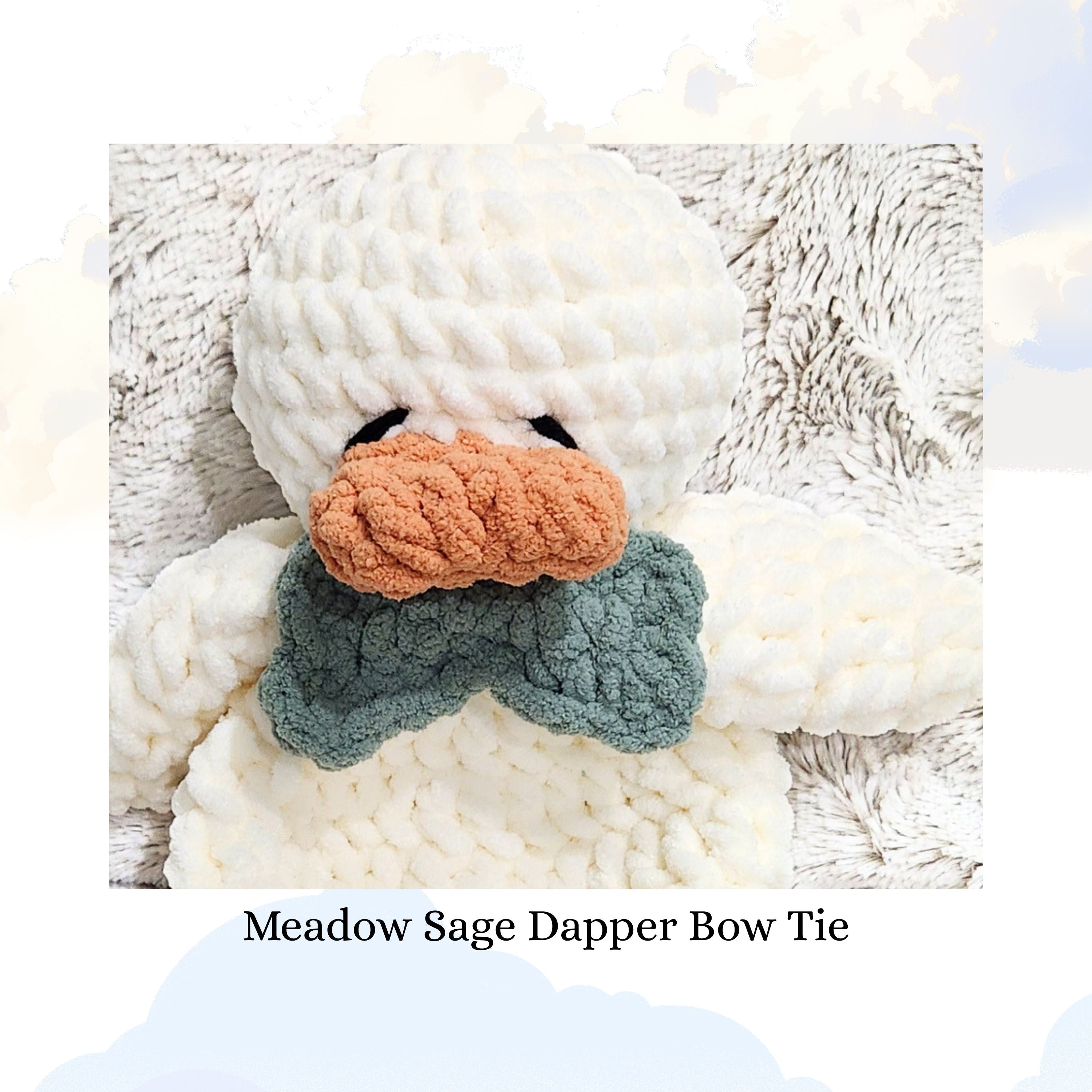 Close up of a Ducky Dreams duck snuggler wearing a dapper meadow sage green bow tie.  The duck is resting on a plush blanket against a whimsical background of soft focus clouds, highlighting the bow tie's crochet texture