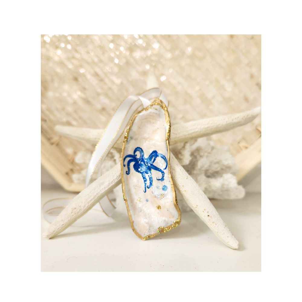 A handcrafted gilded oyster shell ornament featuring a nautical blue octopus design accented with subtle gold sparkle. The shell is finished with a brilliant gold leaf border and a crisp white satin ribbon with gold trim, beautifully propped on a natural starfish and white coral background