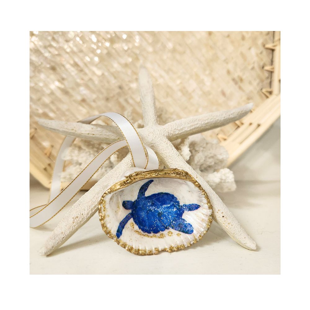A handcrafted gilded ornament made from a unique, deep-cupped cockle shell featuring a nautical blue swimming sea turtle design with a touch of sparkle. The shell is finished with a brilliant gold leaf border and a white satin ribbon with gold trim, beautifully staged on a natural starfish and propped by white coral