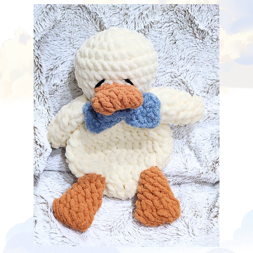A Ducky Dreams vanilla cream crochet duck snuggler wearing an ocean mist blue dapper bow tie. The full body shot shows the duck nestled on a plush blanket set against a whimsical background of soft, dreamy clouds