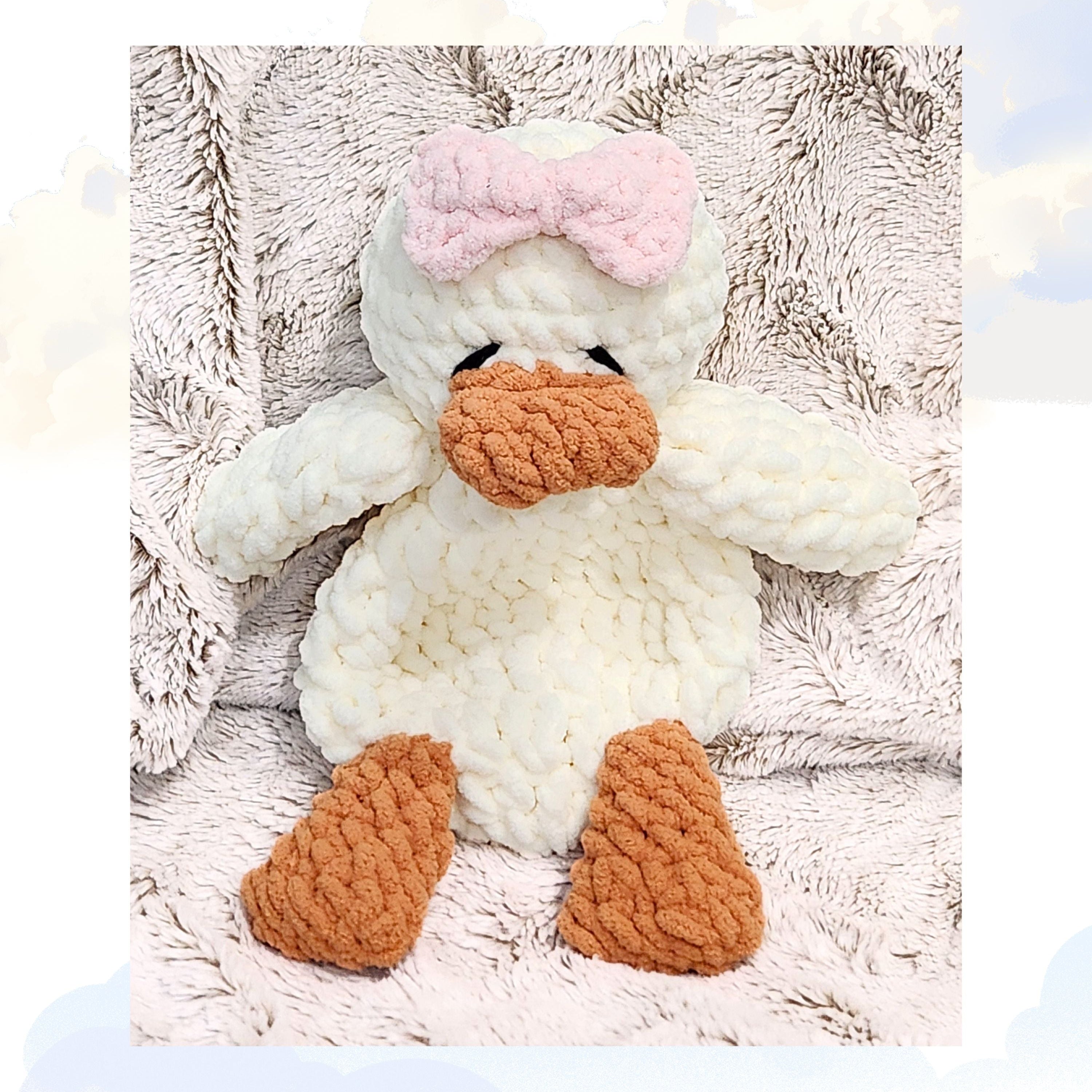 A Ducky Dreams vanilla cream handmade duck snuggler wearing a soft petal pink bow.  The duck is tucked into a plush blanket against a background of soft, whimsical clouds, creating a peaceful and dreamy scene