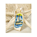 A handcrated gilded oyster shell ornament featuring a sailboat design with white sails trimmed in vivid lime green, highlighted with a delicate touch of sparkle. Finished with a white and gold satin ribbon hanger against a starfish and white coral