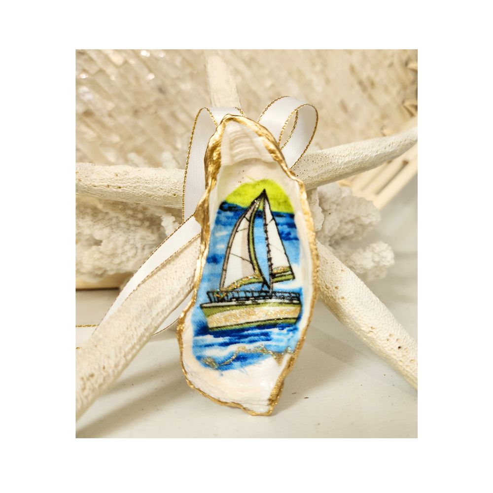 A handcrated gilded oyster shell ornament featuring a sailboat design with white sails trimmed in vivid lime green, highlighted with a delicate touch of sparkle. Finished with a white and gold satin ribbon hanger against a starfish and white coral