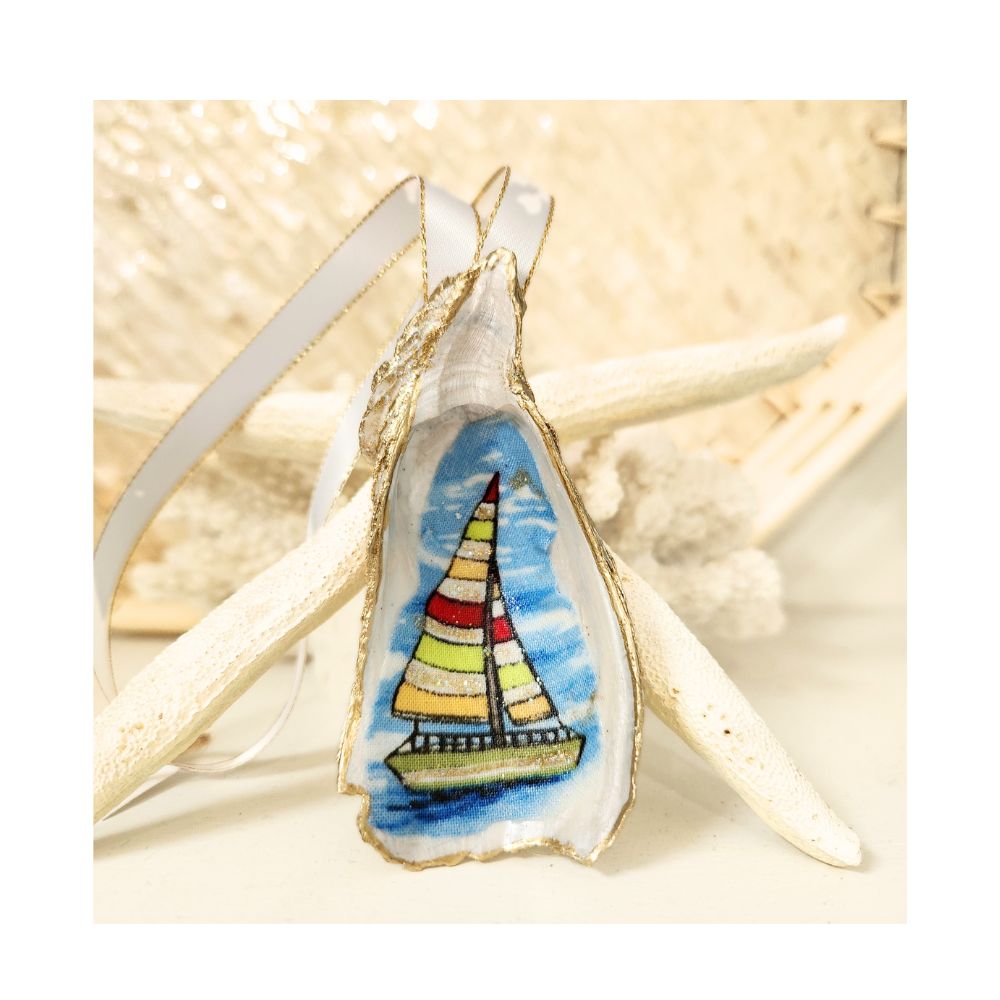 A handcrafted gilded oyster shell ornament featuring a sailboat with vibrant red, white, lime green, and yellow striped sails gliding across blue waters. The design is highlighted with a delicate touch of sparkle and finished with a white and gold satin ribbon hanger against a starfish and white coral