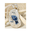 A handcrafted gilded oyster shell ornament featuring a seagull design with an elegant blue and white Chinoiserie floral pattern, finished with a white and gold satin ribbon hanger against a starfish