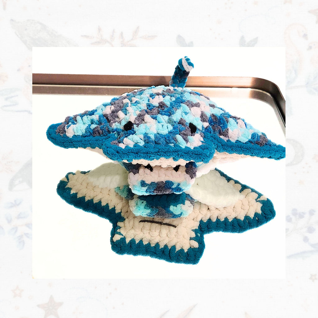A top-down view of the Seraphina and Sol stingray set resting on a mirror, reflecting the artisan crochet underbelly and mouth detail of the mother while the baby ray stays tucked in her signture pouch, by Coastline Boutique.