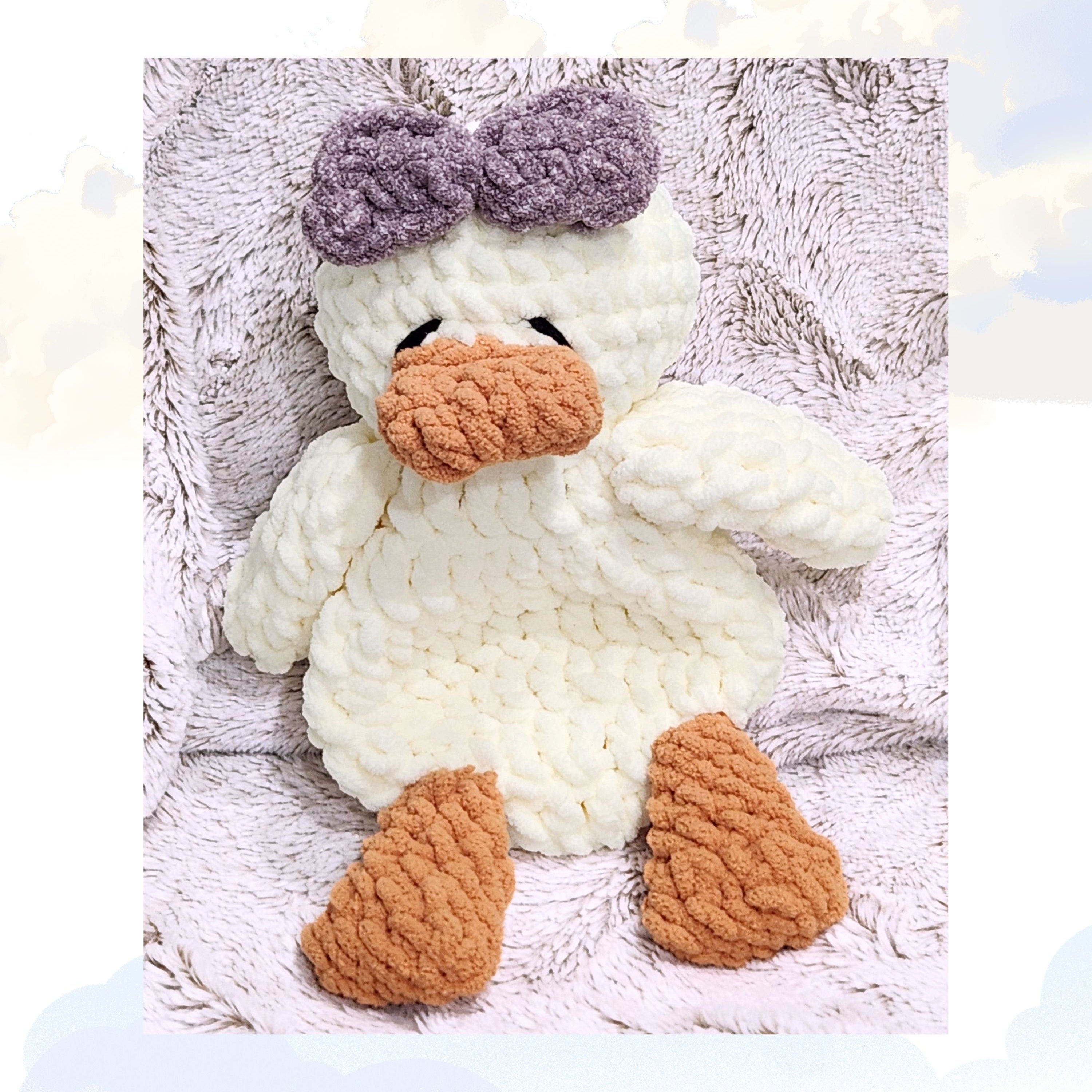 A Ducky Dreams vanilla cream crochet duck snuggler adorned with a sweet lavender purple bow.  The duck is nestled on a plush blanket with a whimsical, soft focus cloud background for a dreamy and calming atmosphere