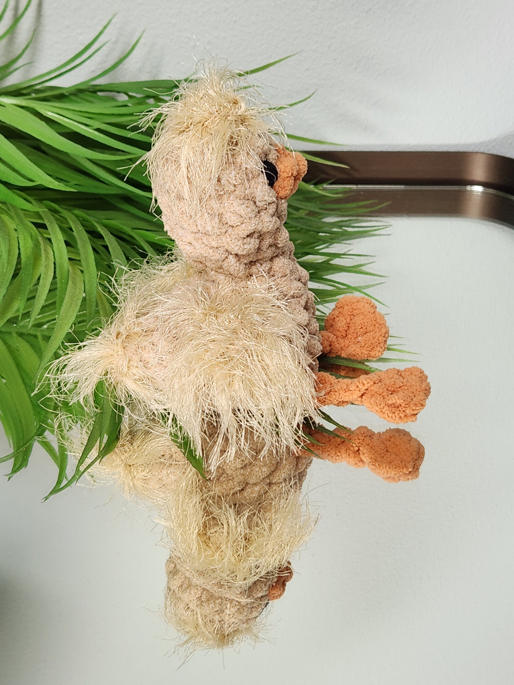 Side profile of Nugget the Gosling on a mirror highlighting the textured fur stitch wings and handcrafted legs in a soft golden yarn