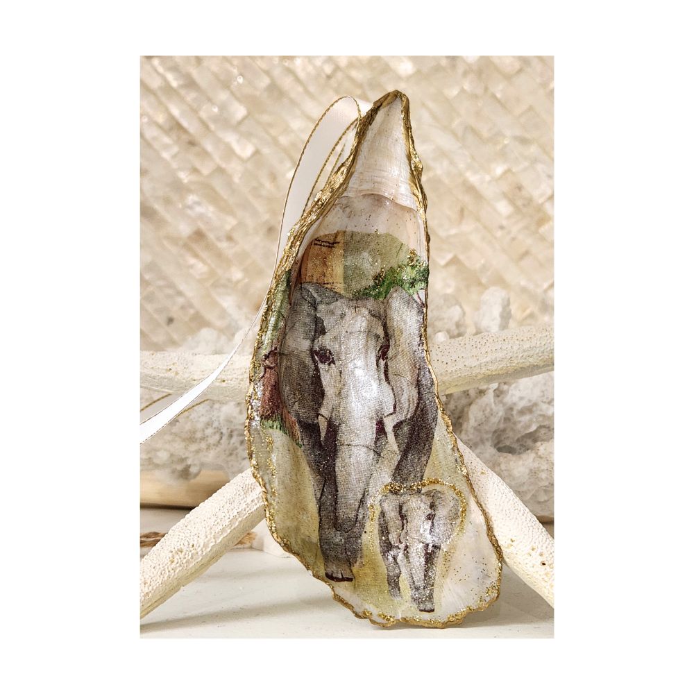 A handcrafted tall gilded oyster shell ornament featuring a front facing mama and baby African elephant design with subtle gold sparkle and green tree details. Staged on a signature background of white coral and a starfish, finished with a white and gold trimmed satin ribbon. 