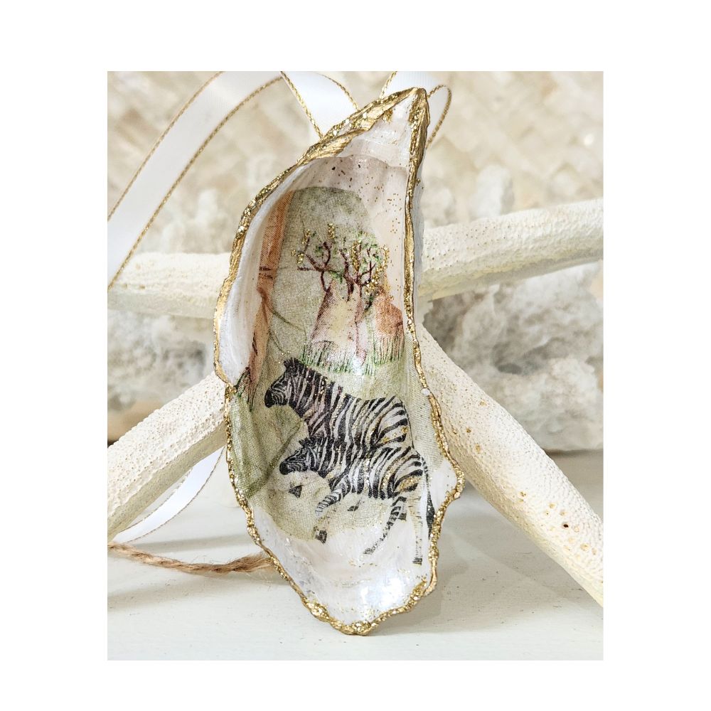 A handcrafted tall gilded oyster shell ornament featuring a design of two zebras with a green grass and safari tree line background. Staged on a signature backdrop of white coral and a starfish, finished with a white and gold trimmed satin ribbon.
