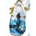 Handcrafted decoupage oyster shell ornament featuring a beautiful mermaid with flowing brown hair overlooking deep blue coastal waters, finished with a tiny starfish at the bottom and a crisp white and gold trimmed ribbon.