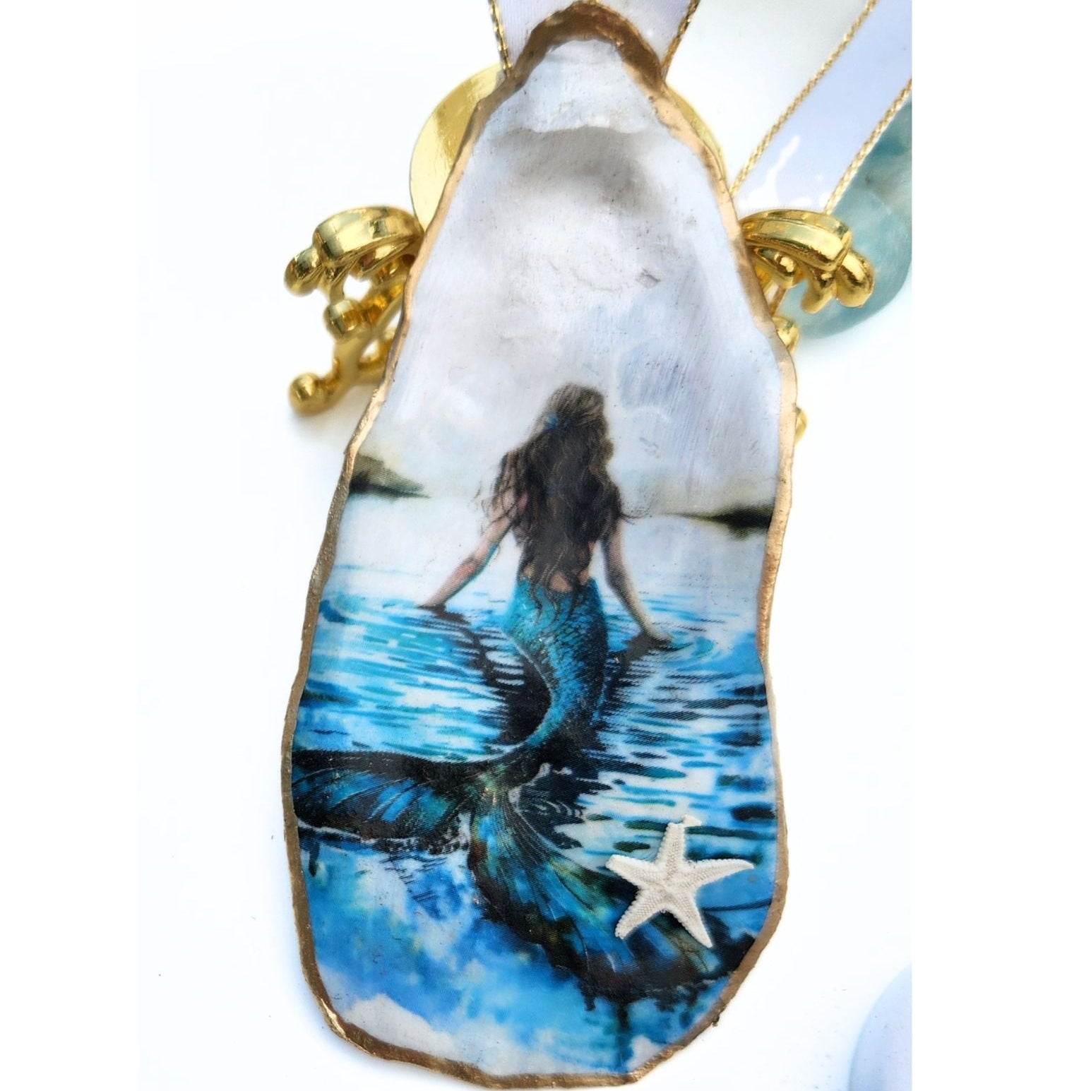 Handcrafted decoupage oyster shell ornament featuring a beautiful mermaid with flowing brown hair overlooking deep blue coastal waters, finished with a tiny starfish at the bottom and a crisp white and gold trimmed ribbon.
